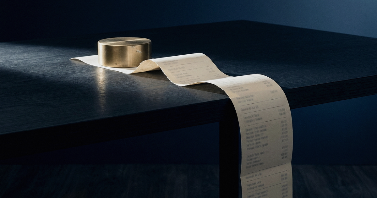 A single continuous ribbon of receipt paper, its near end torn off clean beneath a brass paperweight on a dark desk, running on uninterrupted past the tear, over the desk edge and out of the bottom of the frame.