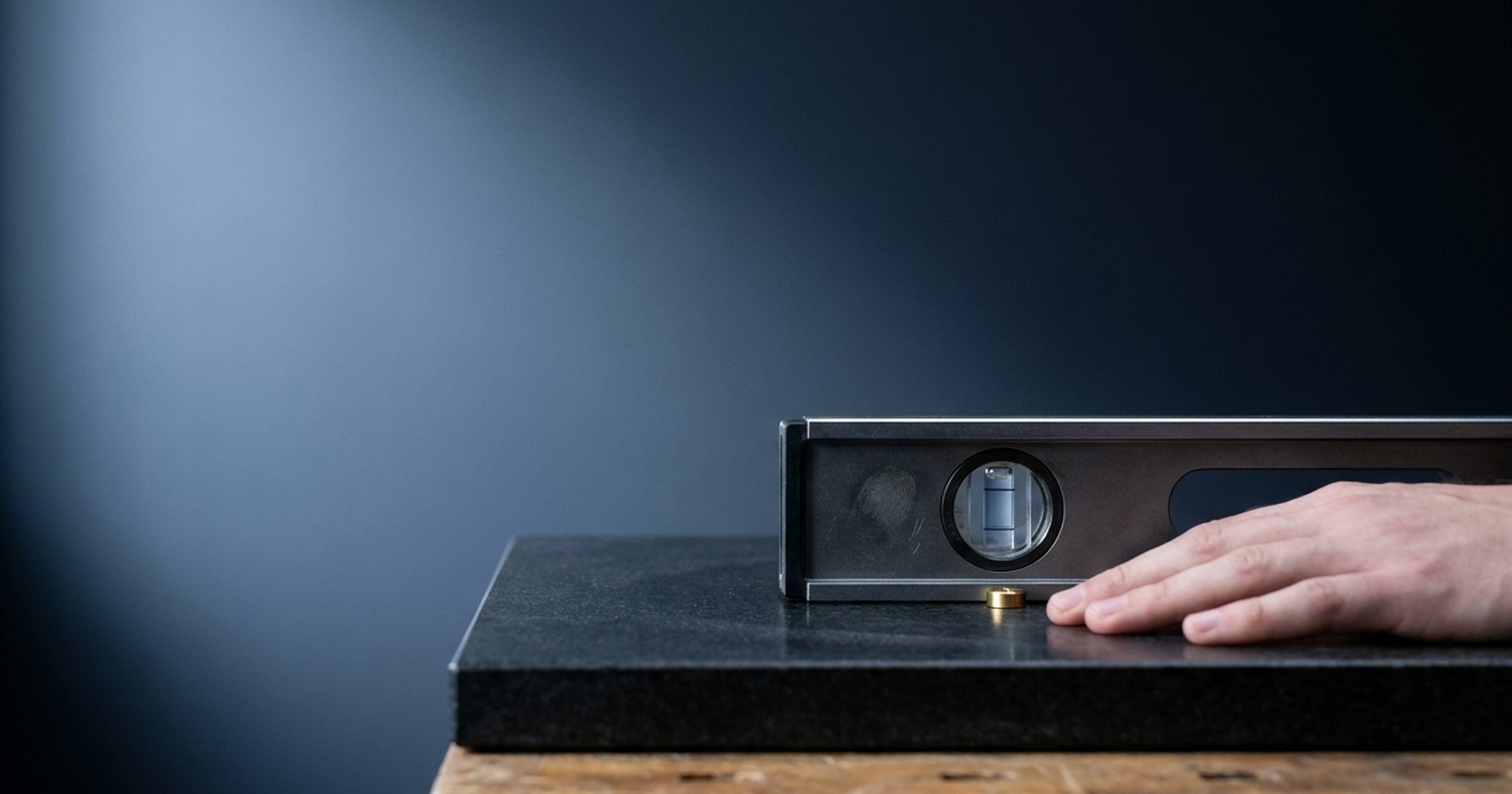 Machinist's level on dark granite, bubble not yet centered, a still hand waiting in soft foreground light.