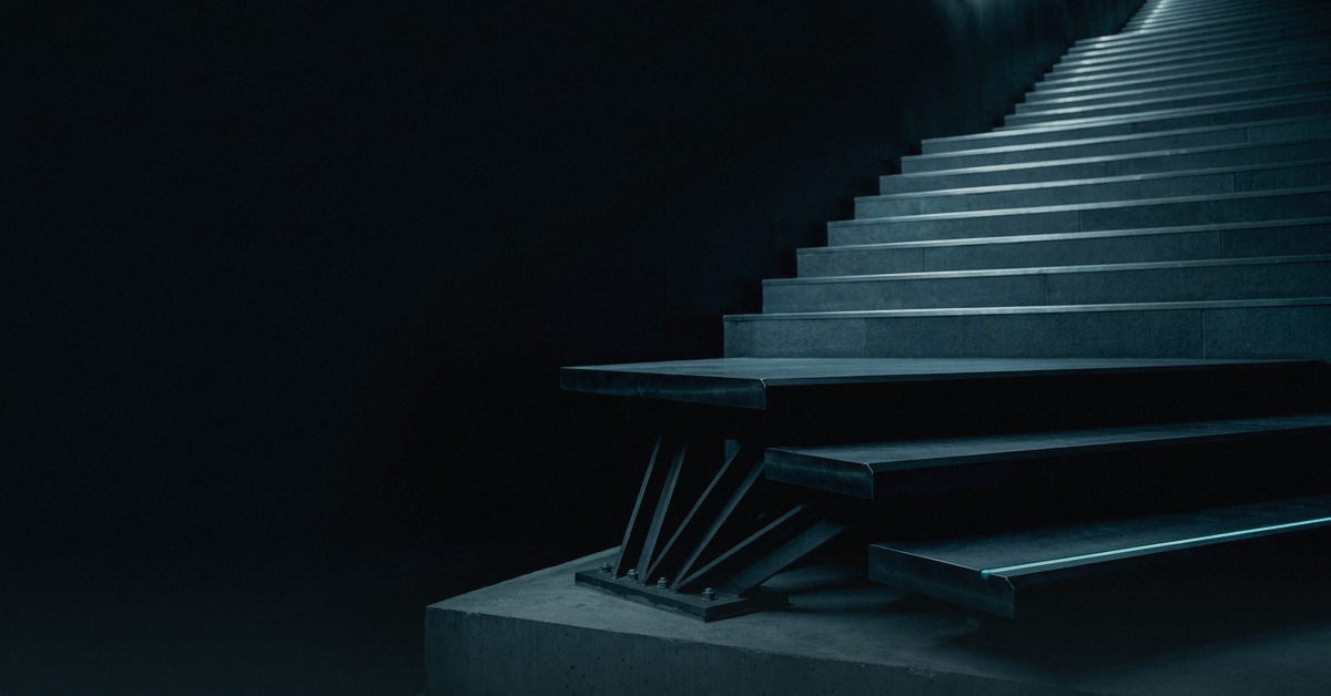 The base of a monumental dark stone staircase photographed at ground level, where the three lowest treads are thick machined steel braced from below by a fan of struts, lit by a single hairline of cold light.