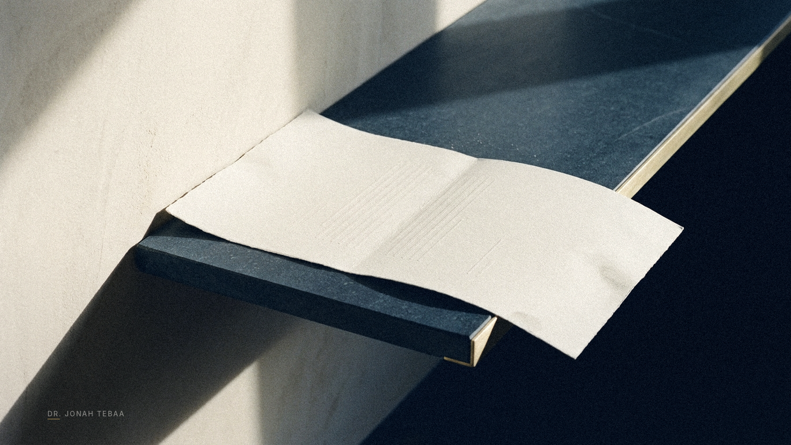 A single sheet of heavy white paper lying on a long dark blue stone table. Its left edge is pressed flush against a pale limestone wall under hard raking light, casting a sharp shadow. Its right edge overhangs a sheared, brass-capped ledge into open darkness, casting no shadow at all.