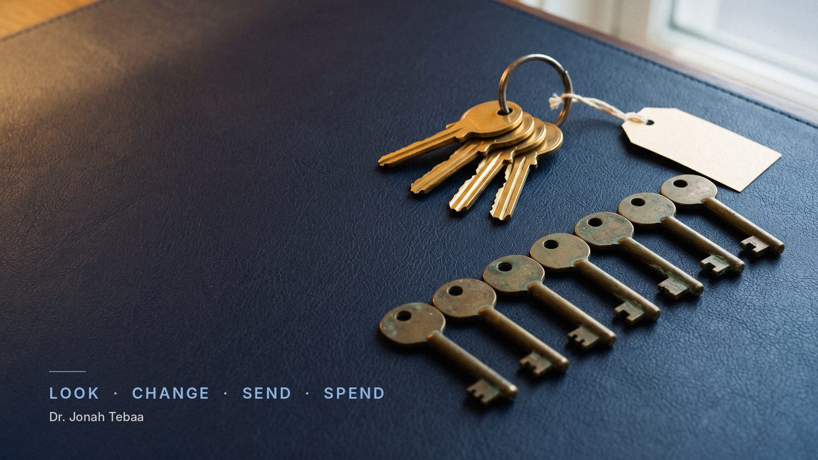 A brass key ring holding four bright, worn keys rests on a navy leather desk blotter in warm lamp light, a small blank tag tied to it. Beside it, seven dull, tarnished keys that have been taken off lie in a neat row. Blue evening light fills the dark office behind. Small text reads 'Look, Change, Send, Spend' and 'Dr. Jonah Tebaa'.