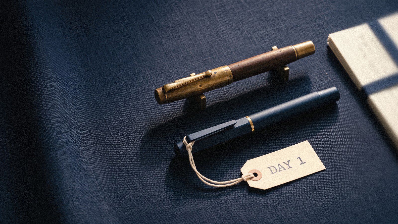 Two fountain pens of a similar shape on navy linen: a worn brass-and-walnut pen raised on small brass rests, and a new matte navy pen with a paper tag reading Day 1 lying in its shadow, beside a bundle of index cards.