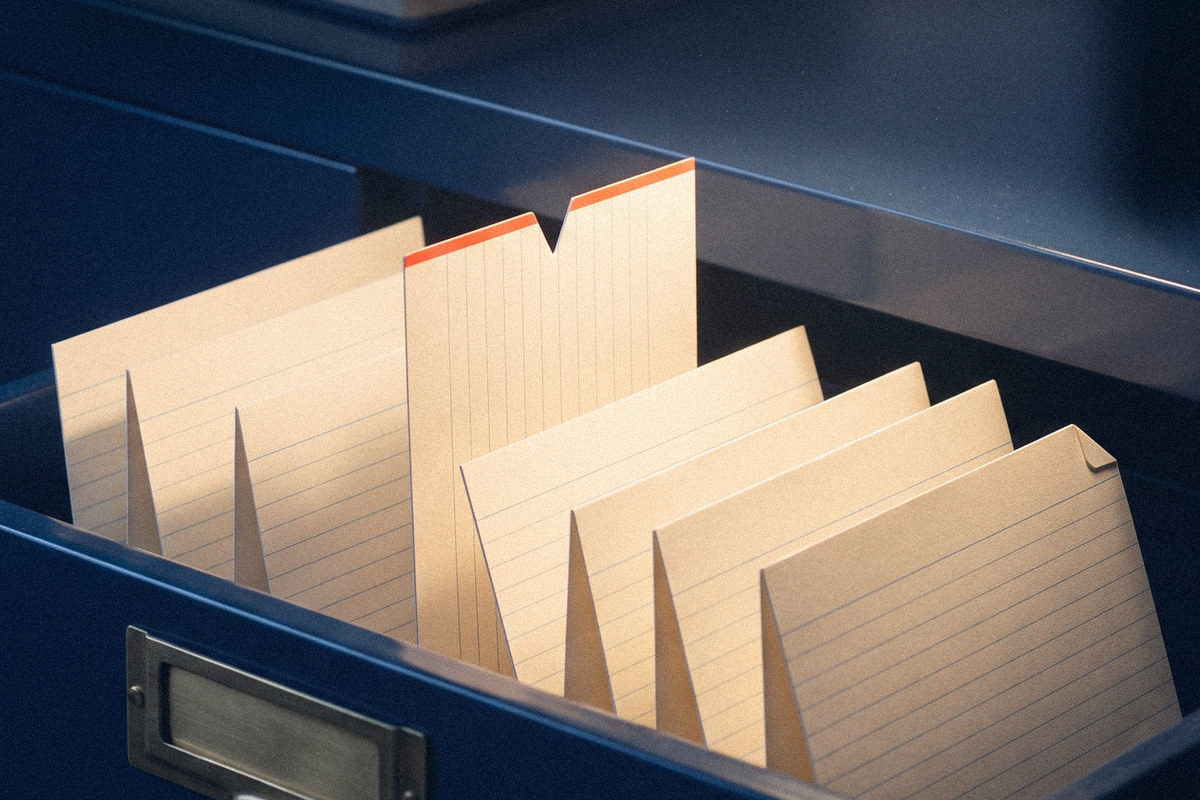 A navy flat-file drawer with one cream index card raised above the others, a small red-orange notch cut in its top edge, in warm desk light.